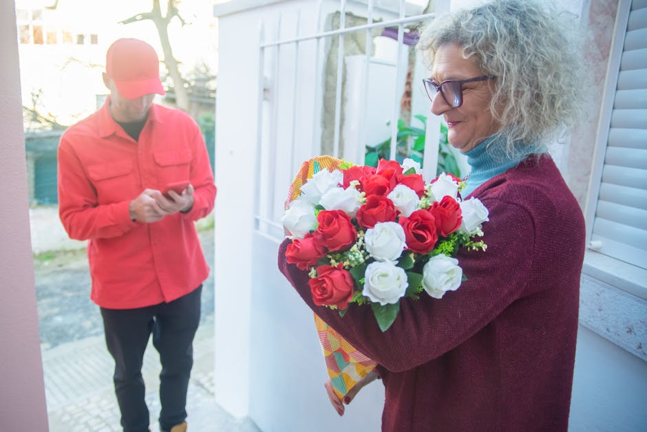 A cheerful woman with curly gray hair, wearing glasses and a burgundy sweater over a blue turtleneck, holds a vibrant bouquet of fresh red and white roses wrapped in colorful patterned paper, engaging