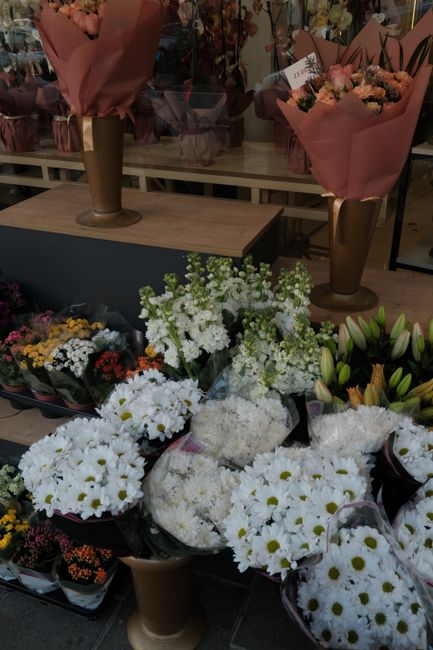 A display of fresh floral bouquets and arrangements at a flower shop, featuring predominantly white daisies with yellow centers, lush white stock flowers, and various other colorful blooms. The flower