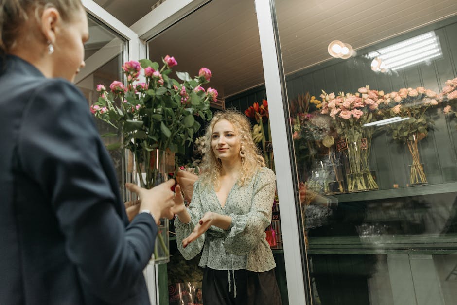 A young woman with curly blonde hair and fair skin receives a bouquet of pink roses from a flower delivery service at the entrance of a flower shop. The bouquet is wrapped in clear cellophane, with vi