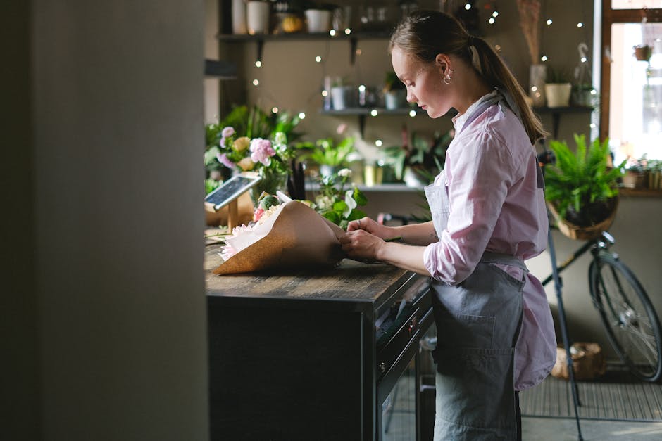 A florist wearing a light pink shirt and grey apron arranging a bouquet of fresh flowers on a wooden counter inside a cozy flower shop. The bouquet includes pink and white blossoms wrapped in brown pa