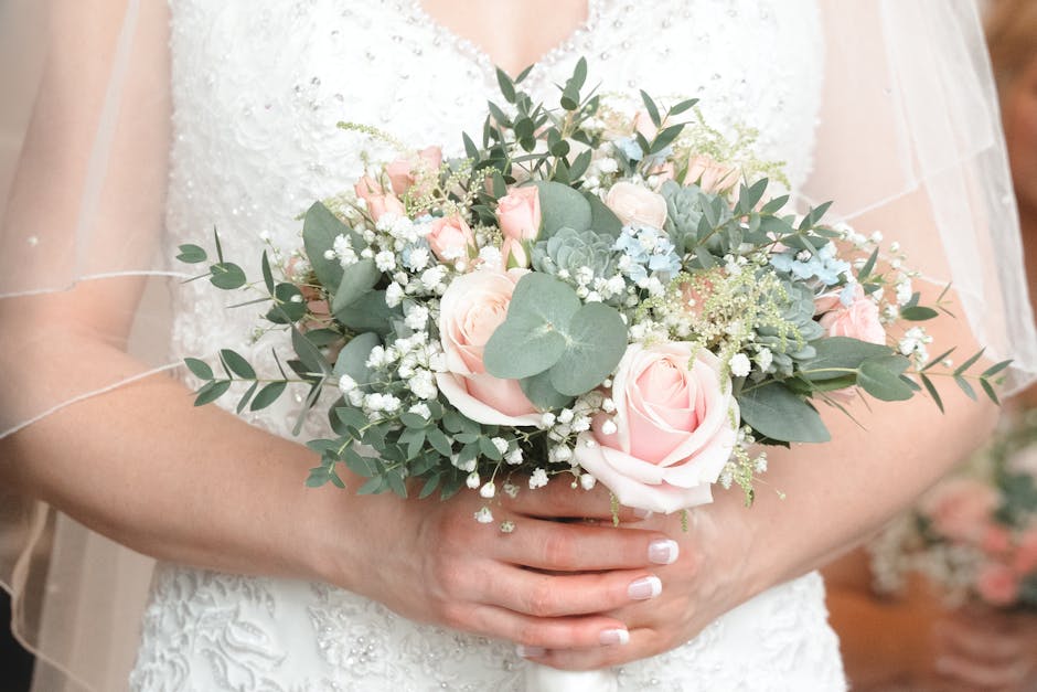 A bride holding a floral bouquet arrangement featuring soft pink roses, white gypsophila, eucalyptus leaves, and pale blue succulents. The bouquet is wrapped in elegant white tissue paper with a satin