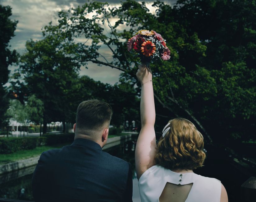 A woman with shoulder-length wavy red hair, wearing a white sleeveless dress, is holding a vibrant bouquet of mixed flowers high in the air with her right hand, while a man in a dark suit stands besid